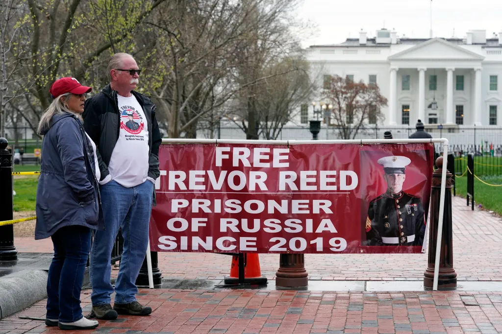 Joey and Paula Reed stand in Lafayette Park next to a banner reading "Free Trevor Reed, Prisoner of Russia Since 2019" with an image of Trevor Reed in his Marine Corps uniform, with the White House in the background.