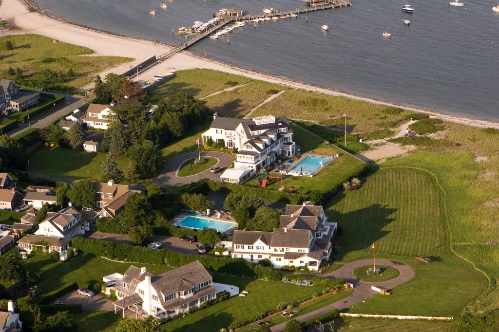 Aerial view of the Kennedy Compound in Hyannisport, Massachusetts, featuring several white houses, green lawns, swimming pools, a sandy beach, and a pier with boats.
