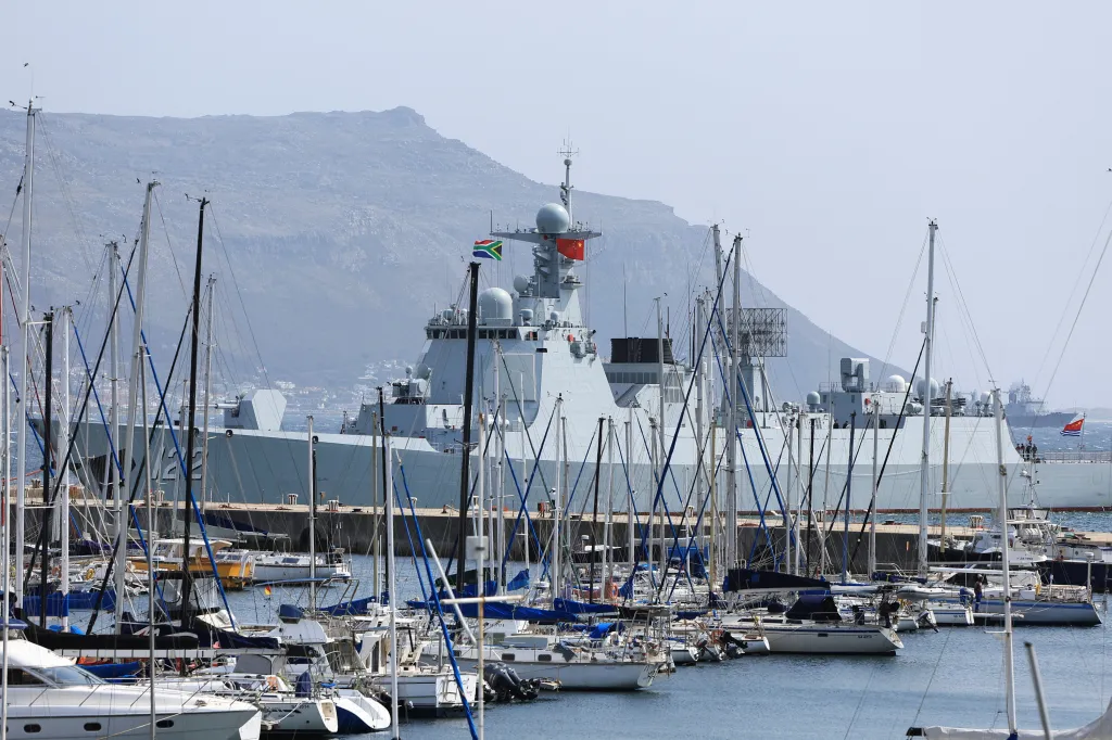 A Chinese naval vessel with a Chinese flag and a South African flag is docked in Simon's Town Naval base, with numerous sailboats in the foreground and a mountain in the background.