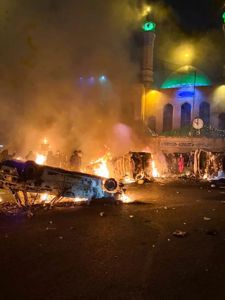 Protests in Tehran with burning vehicles and a mosque in the background.