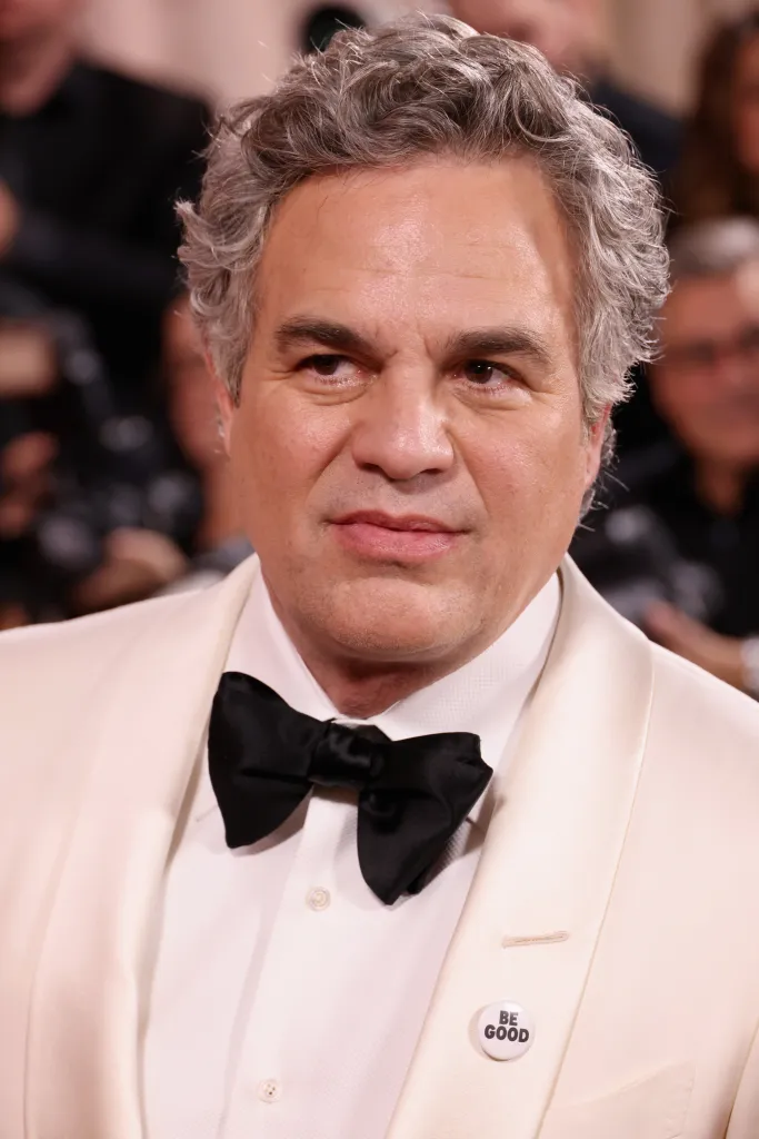 Mark Ruffalo wearing a white tuxedo jacket with a black bow tie and a "BE GOOD" pin on the lapel.