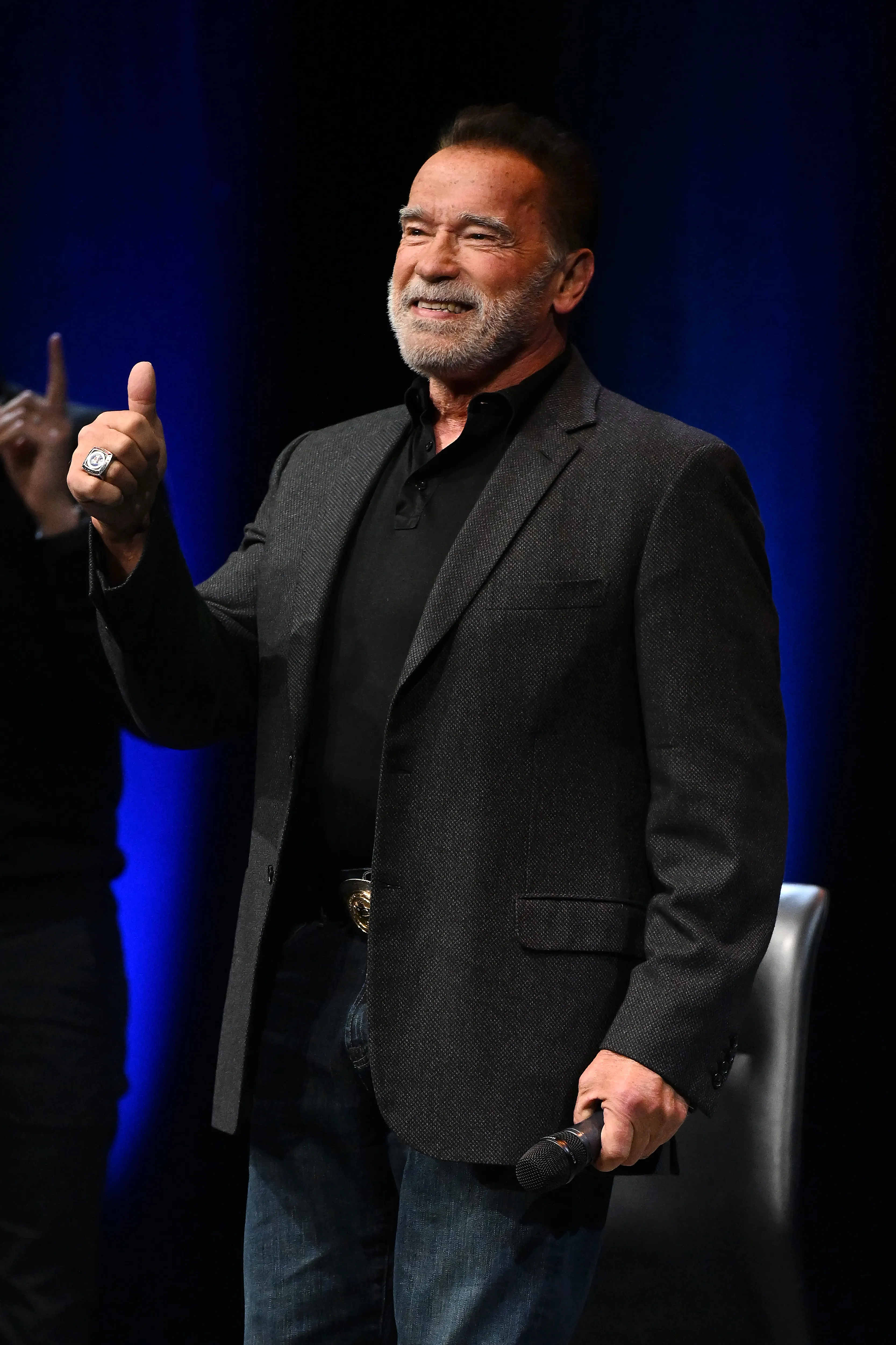 Arnold Schwarzenegger smiling and giving a thumbs up while holding a microphone.