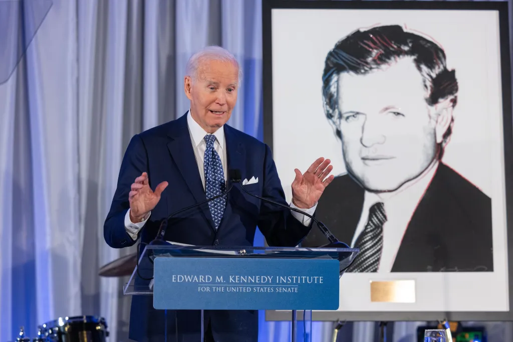 Joe Biden speaking at a podium with the Edward M. Kennedy Institute logo, with a large pop-art style portrait of Edward M. Kennedy behind him.