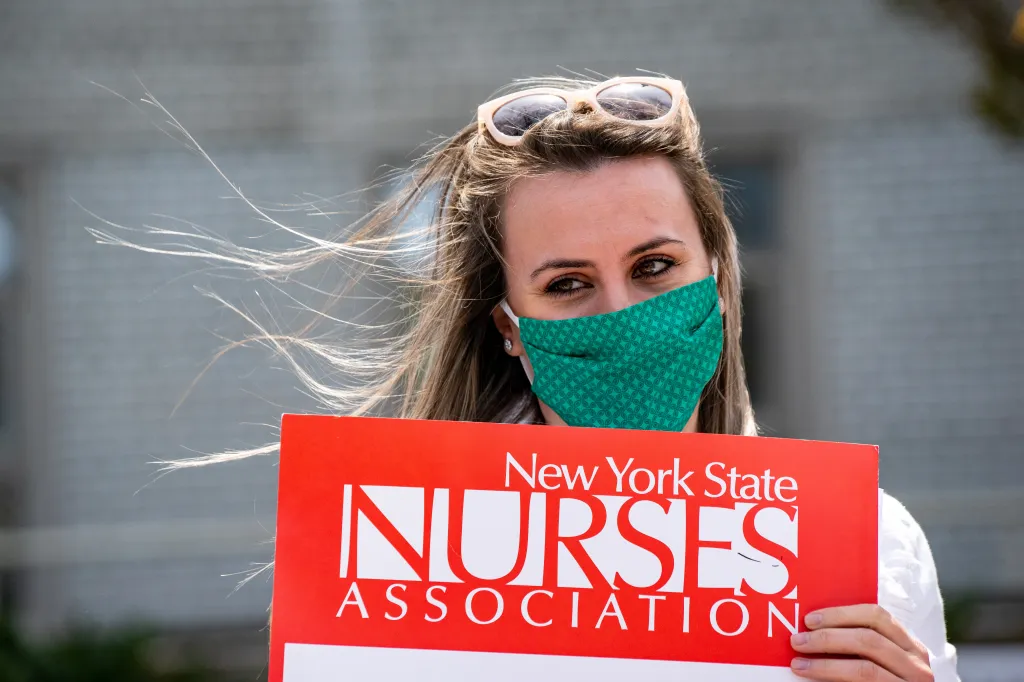 A nurse in a green patterned face mask and sunglasses holds a red sign that reads "New York State Nurses Association."