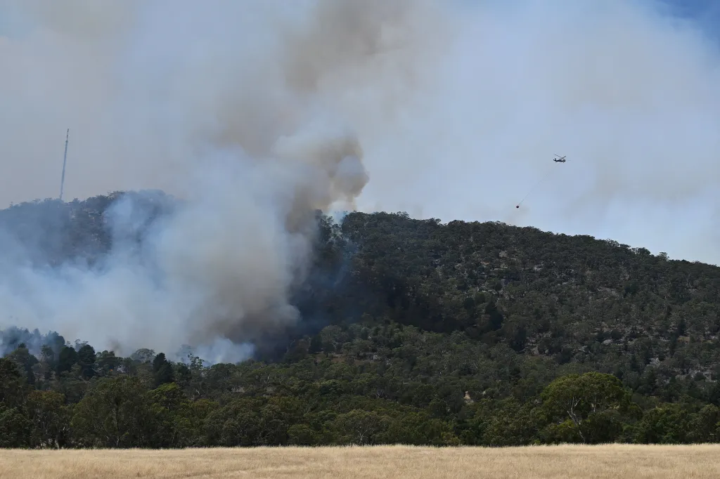 A firefighting helicopter carries a water bucket over a bushfire creating large plumes of smoke over a forested hill.