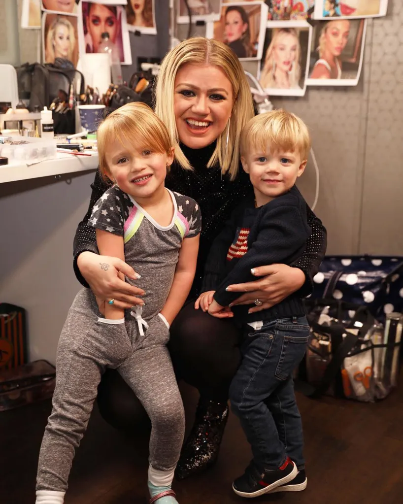 Kelly Clarkson smiling and kneeling between her two young children, a girl and a boy, in a dressing room.