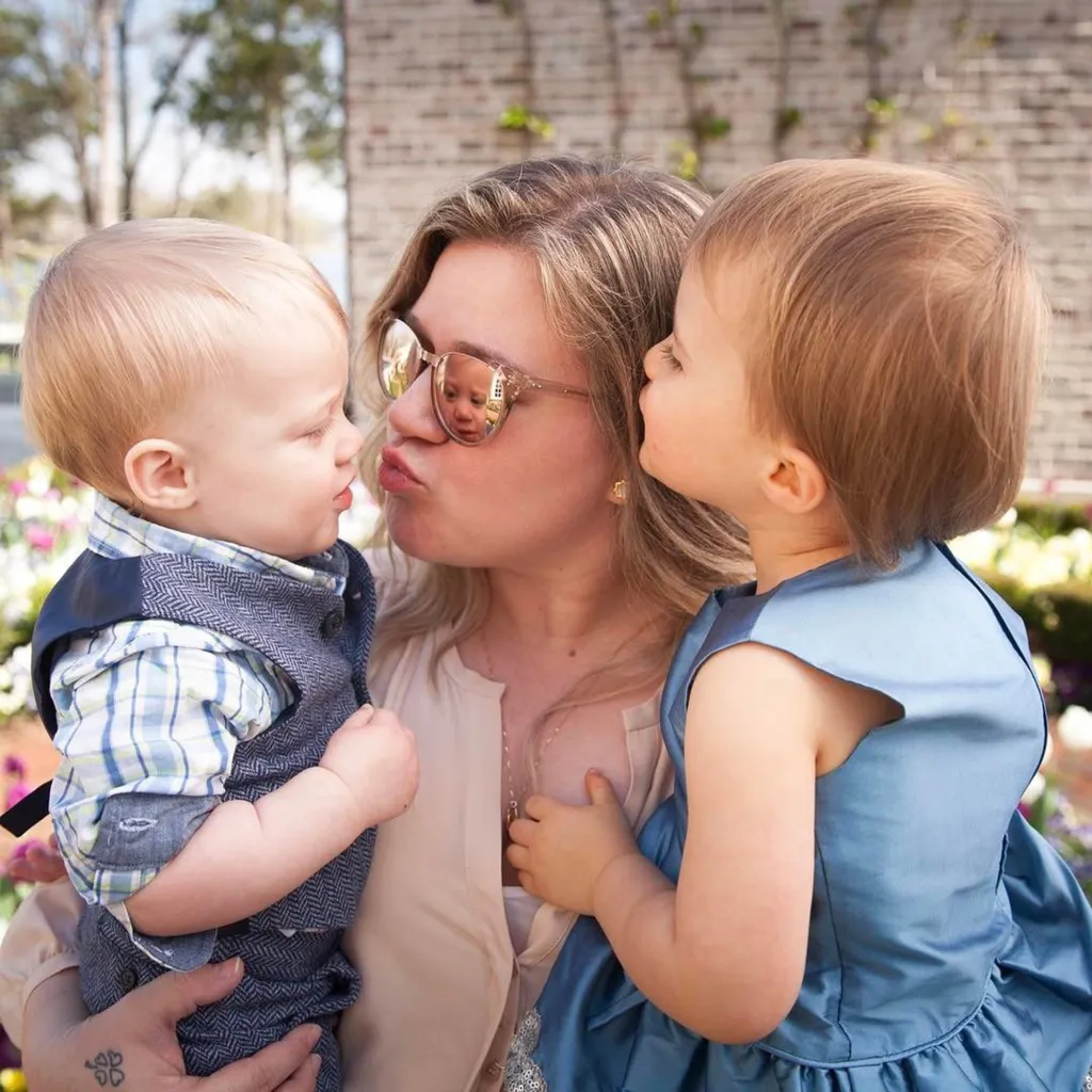 Kelly Clarkson holding her two children, a boy in a vest and a girl in a blue dress.