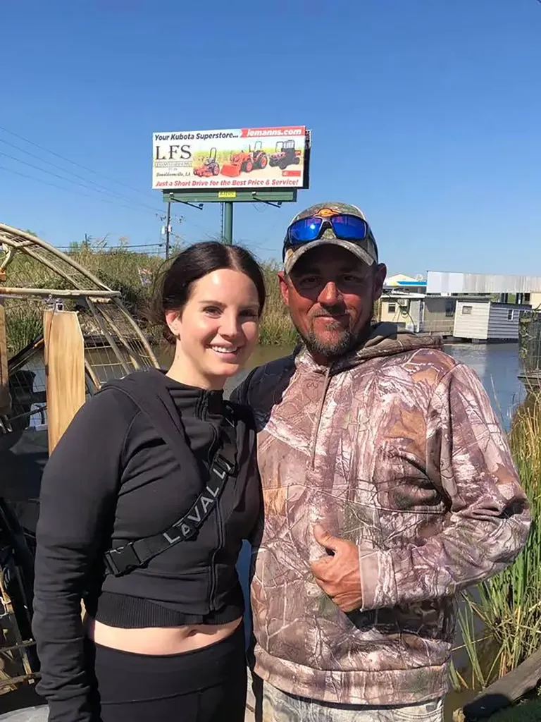 Lana Del Rey standing next to Jeremy Dufrene near a body of water with a billboard in the background.