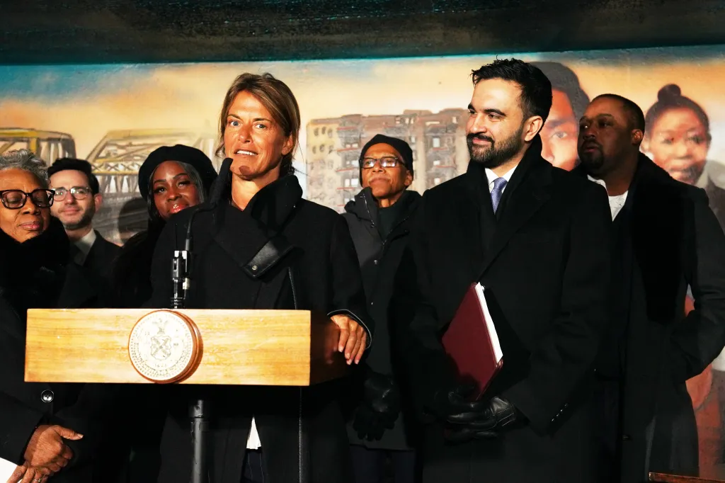 Dina Levy speaking at a podium with Mayor Zohran Mamdani and others standing behind her during a press conference.