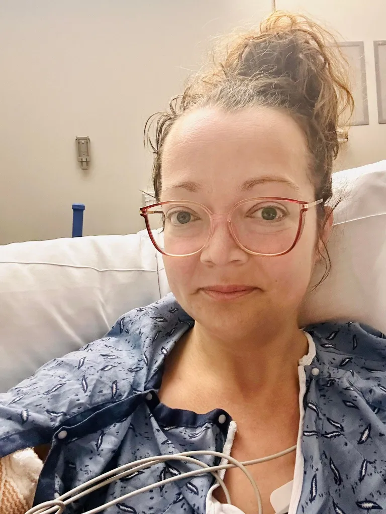 A woman in a hospital gown and glasses is lying in a hospital bed, looking at the camera.
