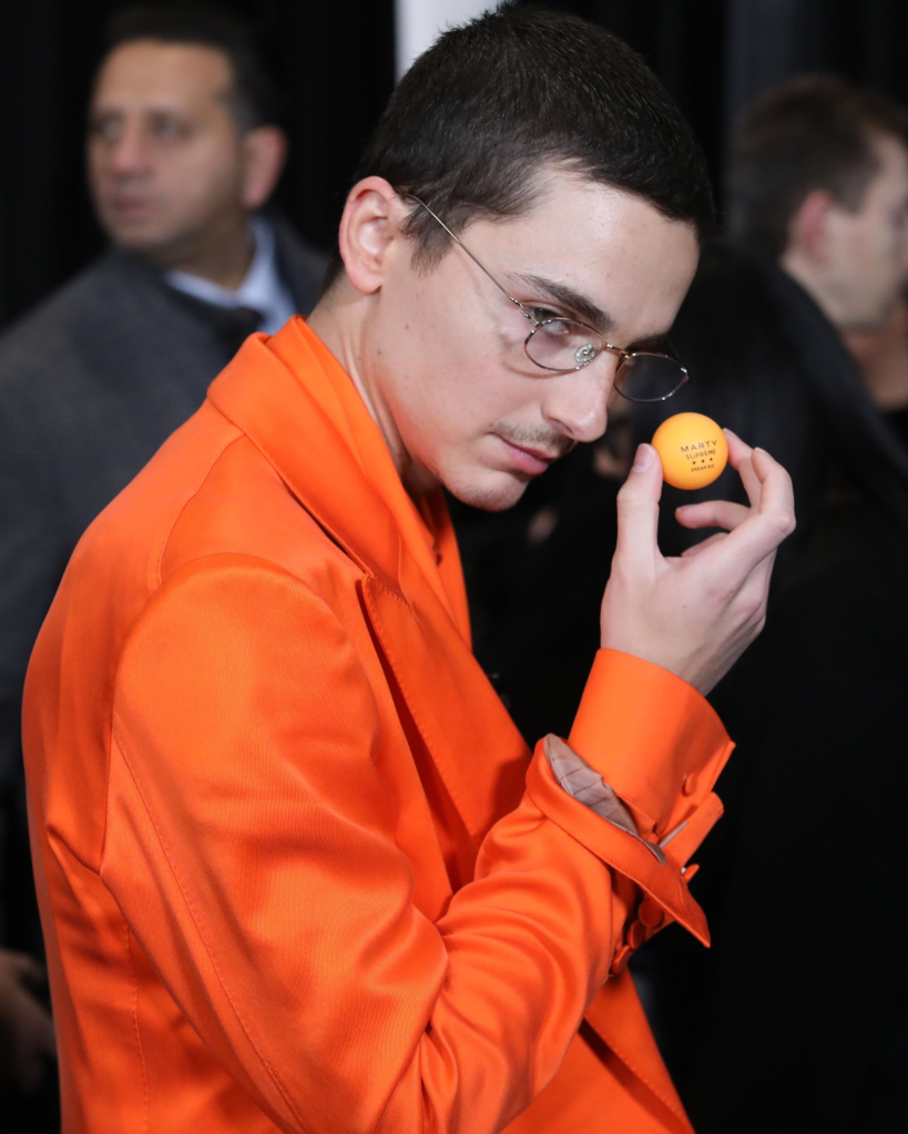 Timothée Chalamet wearing an orange suit, holding an orange ping pong ball with "Marty Supreme Dream Big" printed on it.