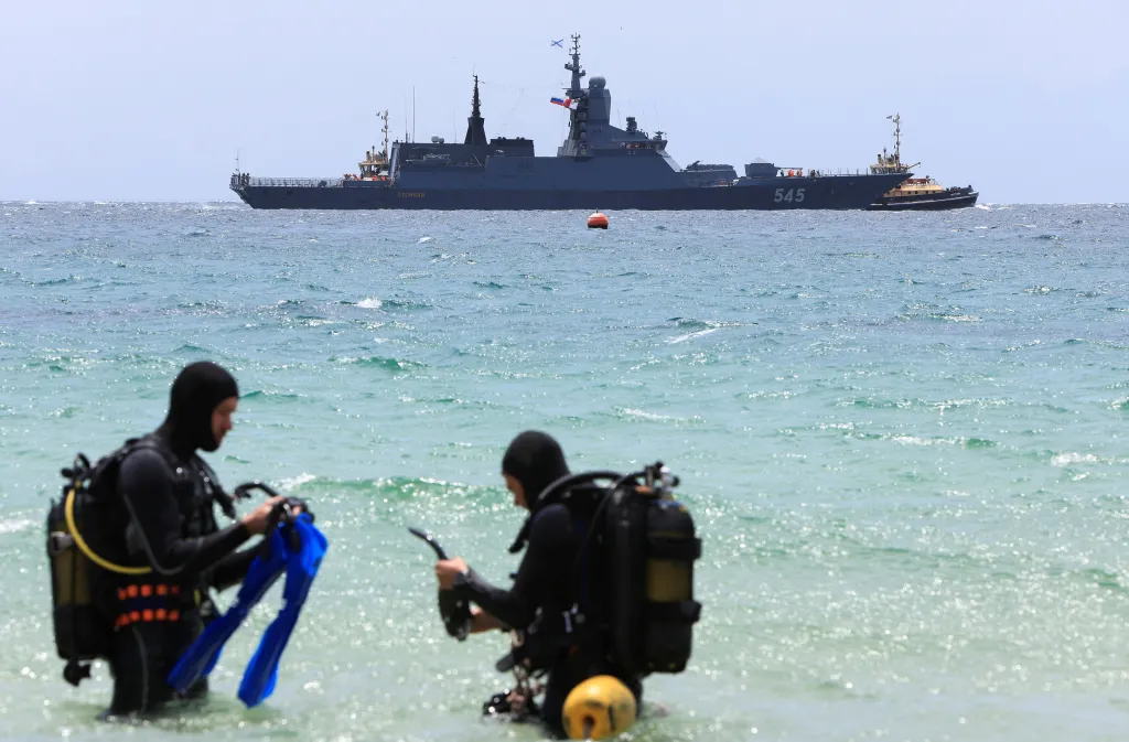 Two divers in wetsuits and an oxygen tank on their backs, stand in the sea in the foreground, with a large grey warship flying the Russian flag visible in the background.