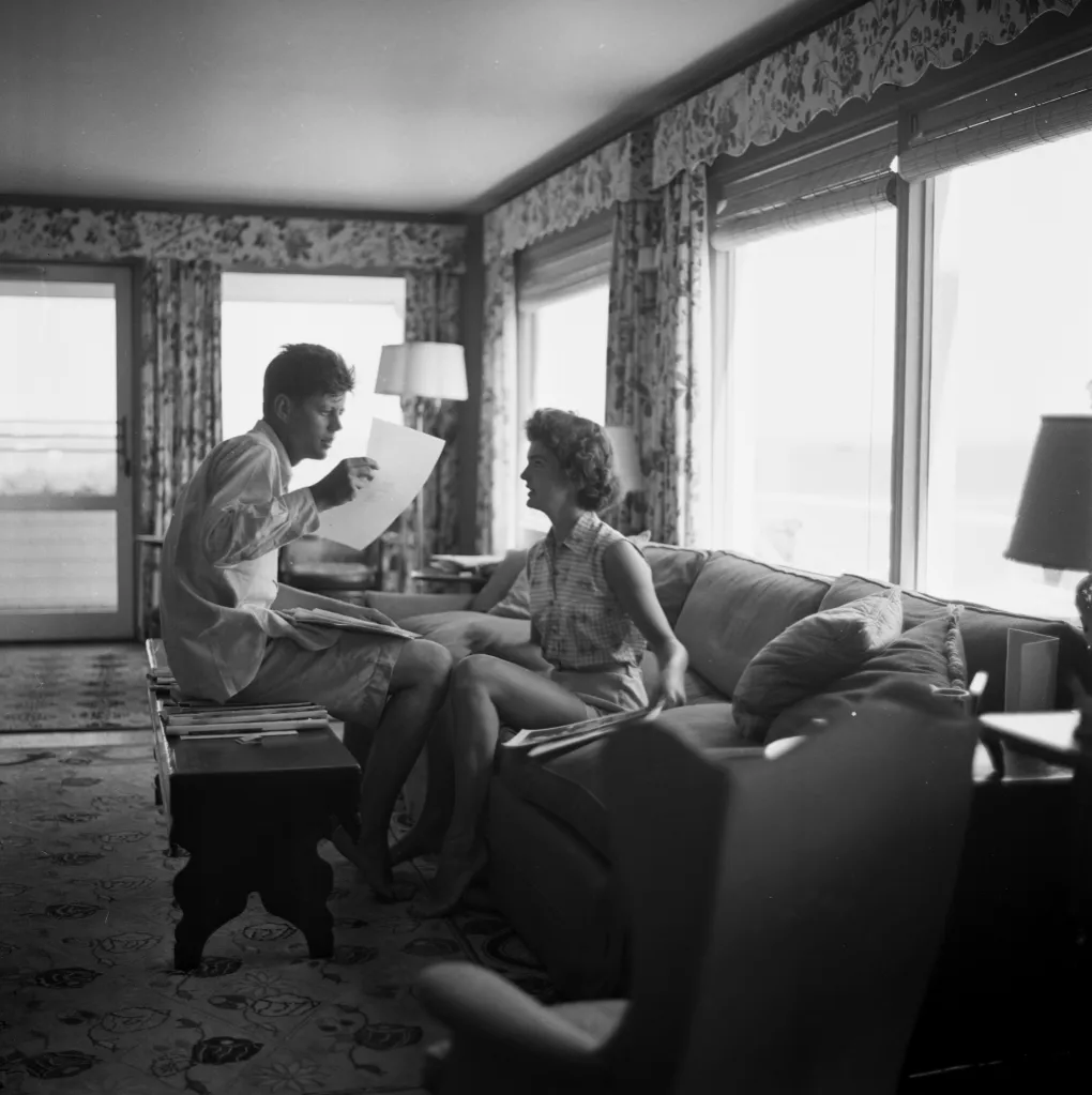 John F. Kennedy and Jacqueline Bouvier review papers on a coffee table.