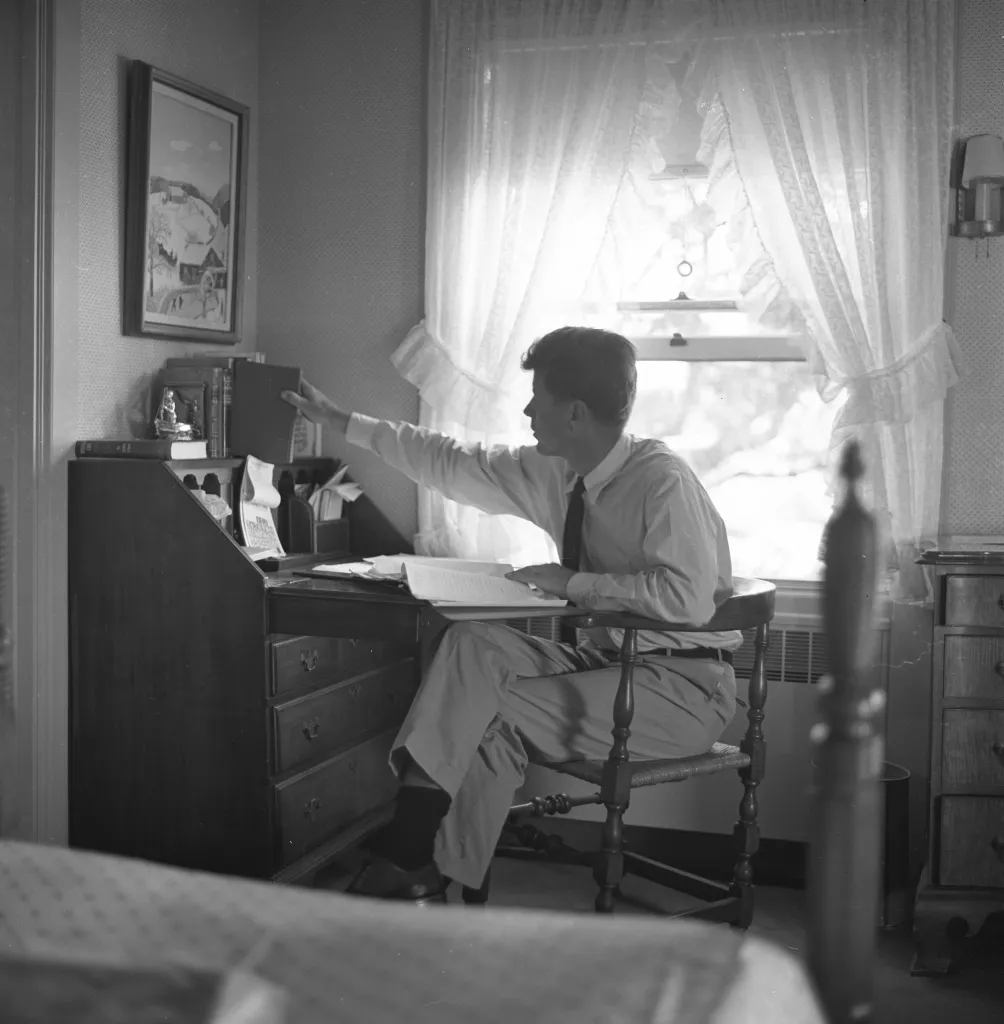 Senator John F. Kennedy reaching for a book while sitting at a desk.