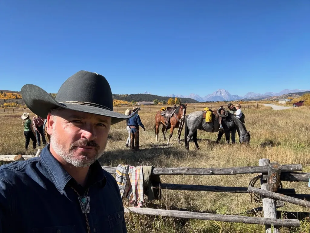 Tom Welling in a cowboy hat and bolo tie, in a field with horses and mountains in the background.
