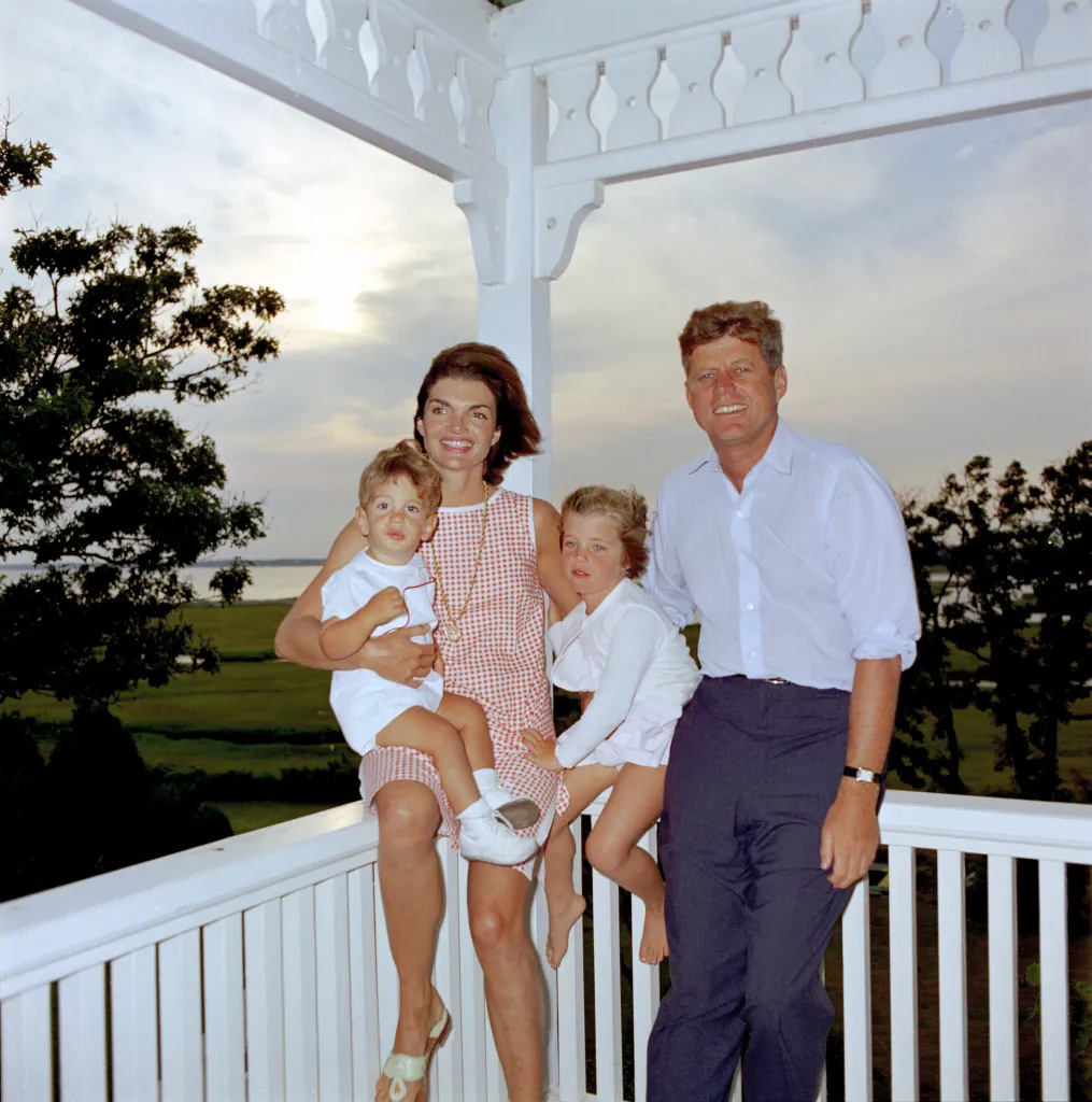 John F. Kennedy and Jacqueline Kennedy with their two young children, John Jr. and Caroline, on a porch overlooking water.