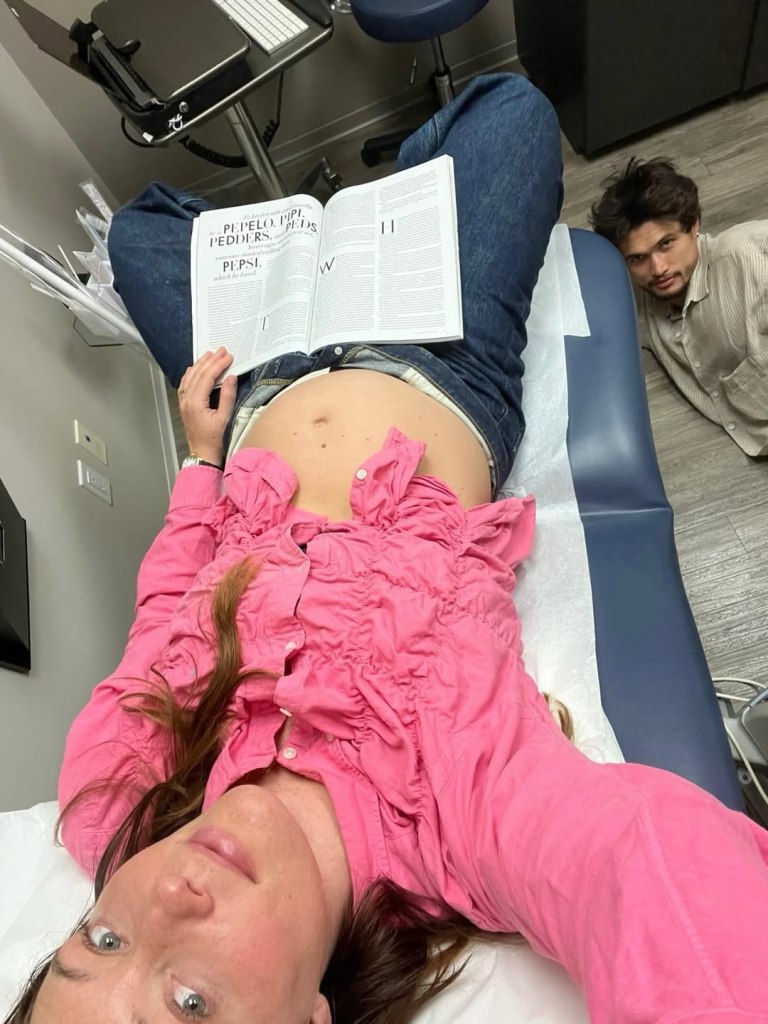 Camille Summers-Valli lying on an examination table with her pregnant belly exposed, reading a book, as Charles Melton looks on.