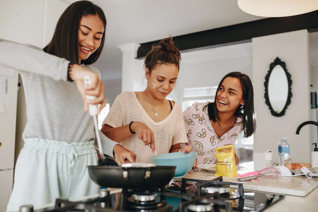 Three smiling girls cooking breakfast together in a kitchen.