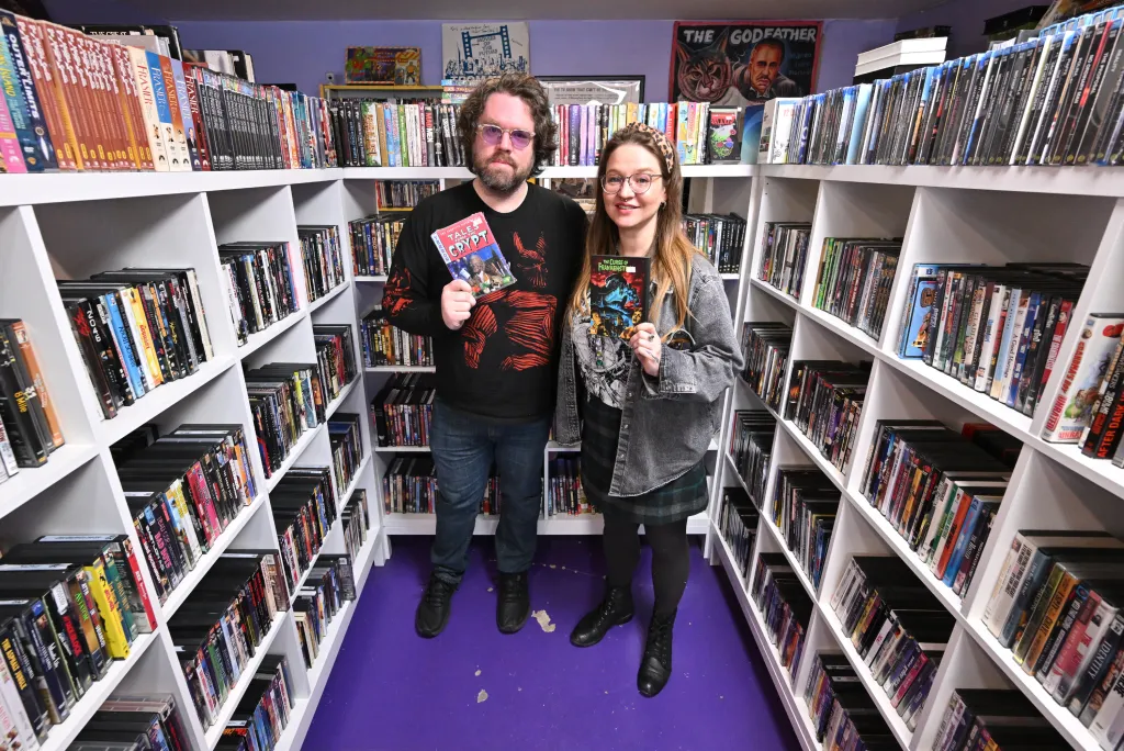 Two people holding DVDs, a man holding "Tales from the Crypt" and a woman holding "The Curse of Frankenstein," standing between shelves of movies in a video store.