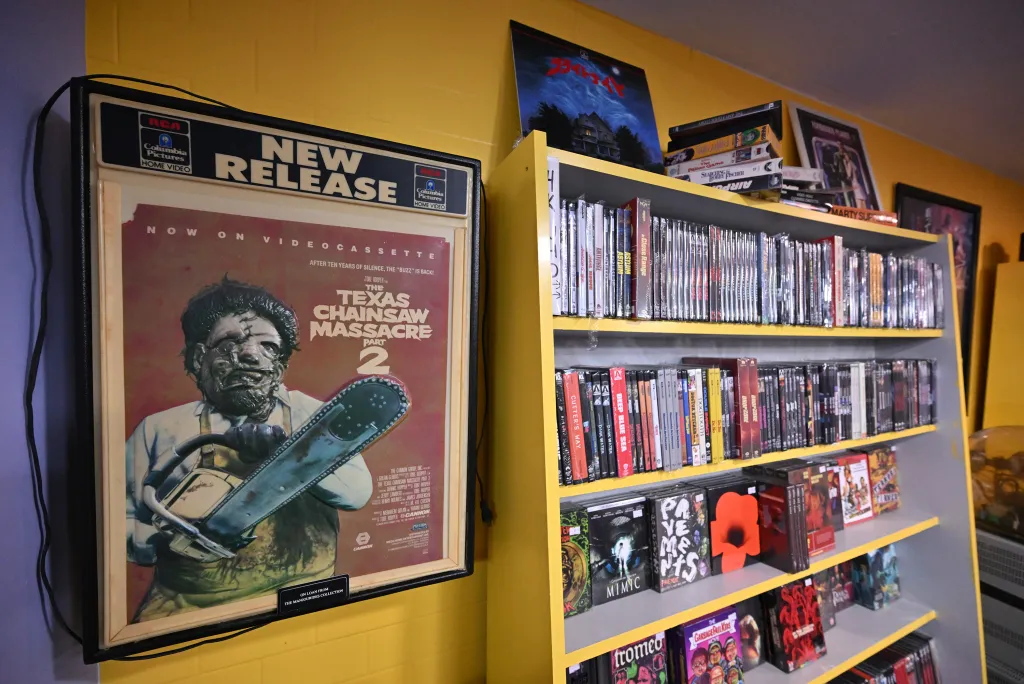 A video store display features a poster for "The Texas Chainsaw Massacre Part 2" and shelves filled with VHS tapes and DVDs.