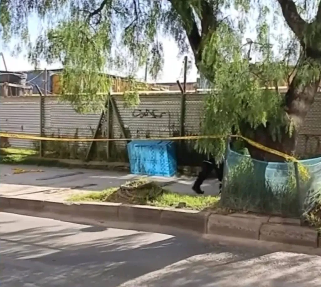 A street scene with a wardrobe covered in blue tarp near a tree and a fence, cordoned off by yellow tape.