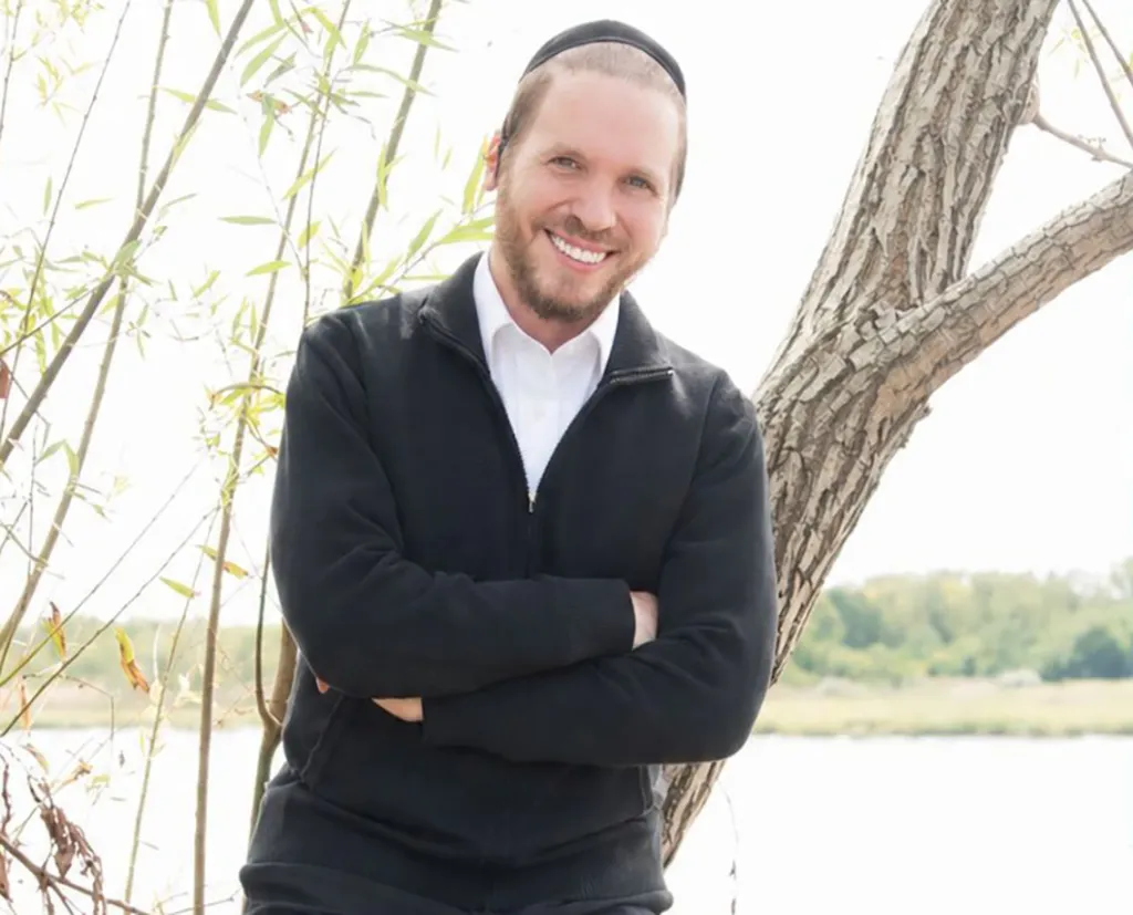 Photo of Rabbi Nuchem Yasir Ebeth smiling, wearing a black yarmulke and black jacket, leaning against a tree.