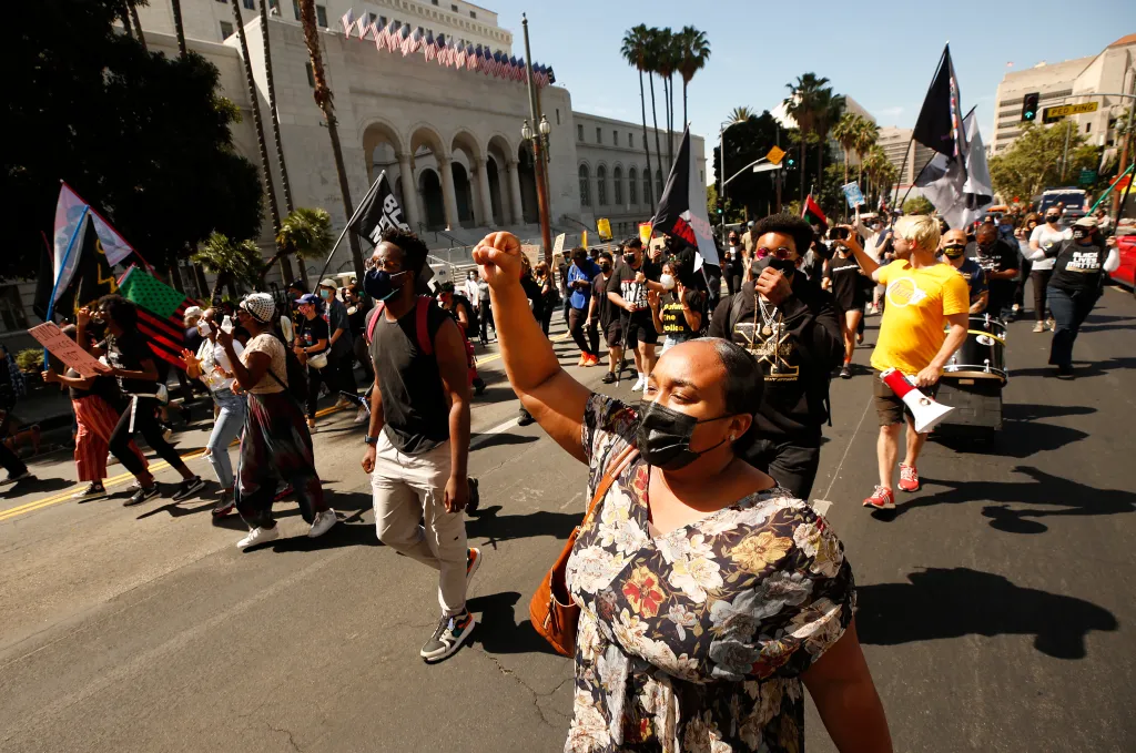 A crowd marches down Spring Street, with a woman in a floral dress and black mask raising her fist in the foreground.