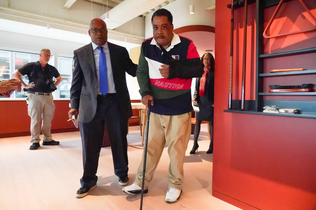 Timothy Brown, injured with his arm in a sling and using a cane, stands next to attorney Derek Sells, both men looking toward the camera.