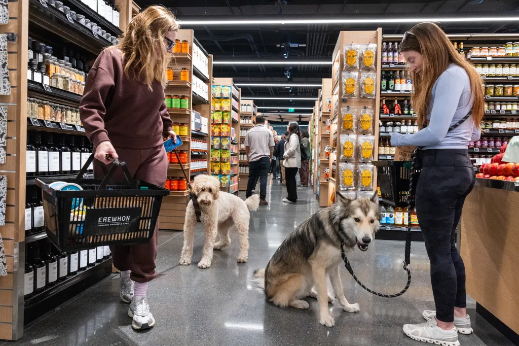 Two women shop in a grocery store with two dogs on leashes.
