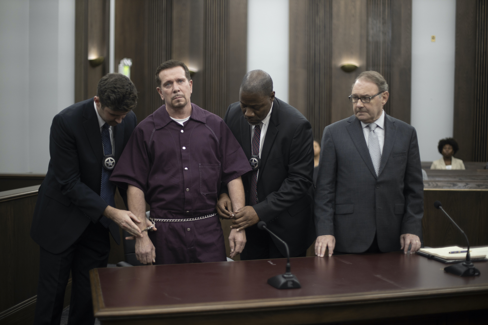 Bobby J Brown in a purple prison uniform stands in a courtroom, chained at the waist and wrists, flanked by two officers and another man in a suit.