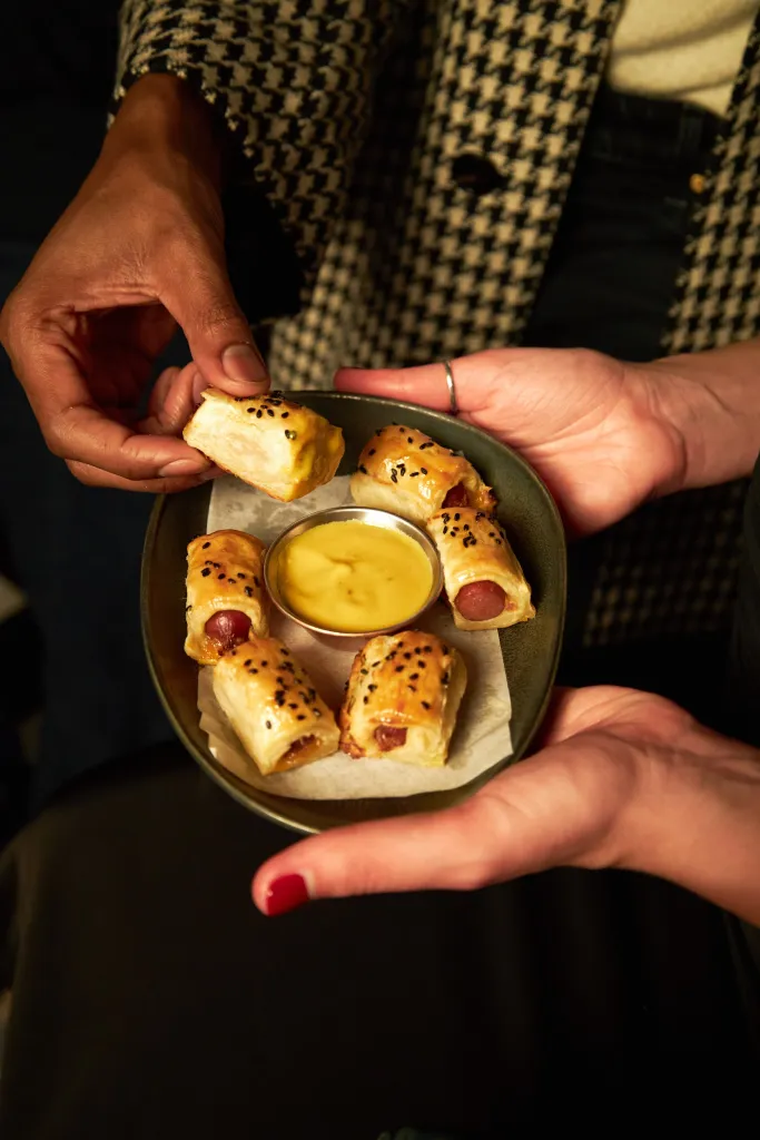 Two hands holding a platter of pigs in a blanket with a mustard dipping sauce.