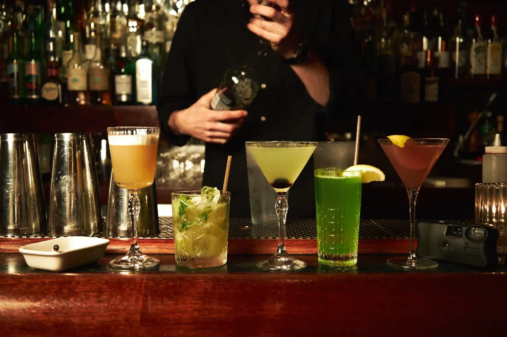 Five cocktails lined up on a dark wooden bar, with a bartender in a black shirt preparing a drink in the background.