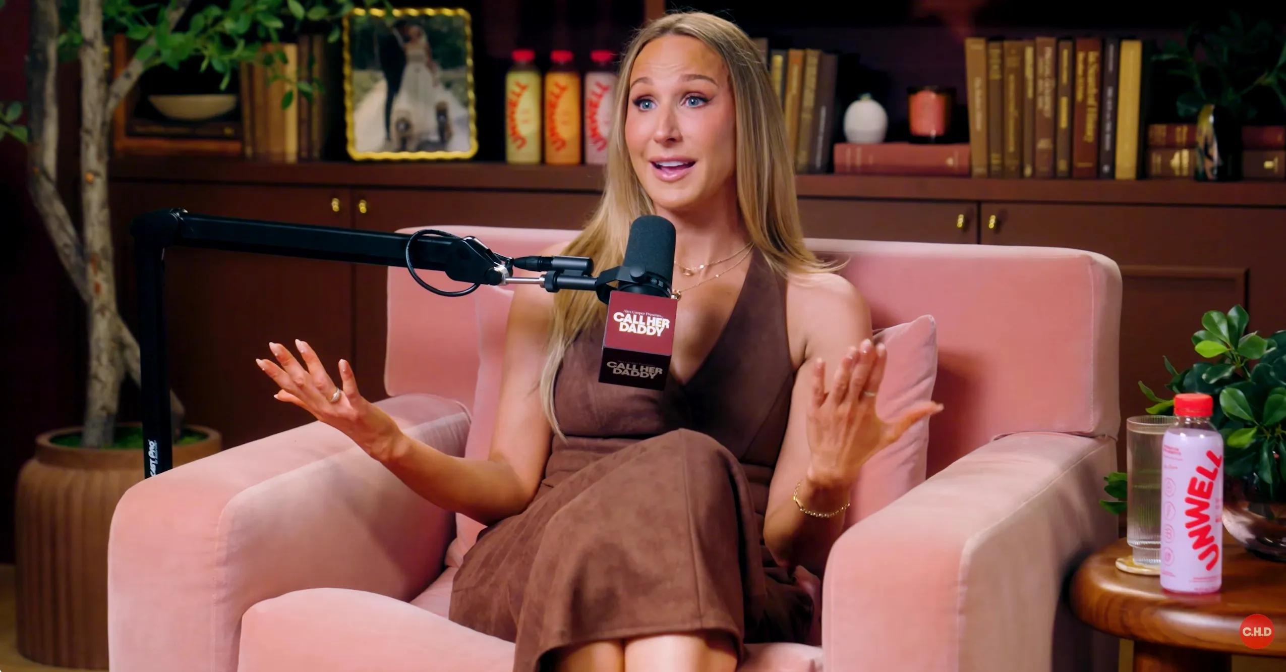 Woman with long blonde hair in a brown dress sits on a pink chair speaking into a microphone.