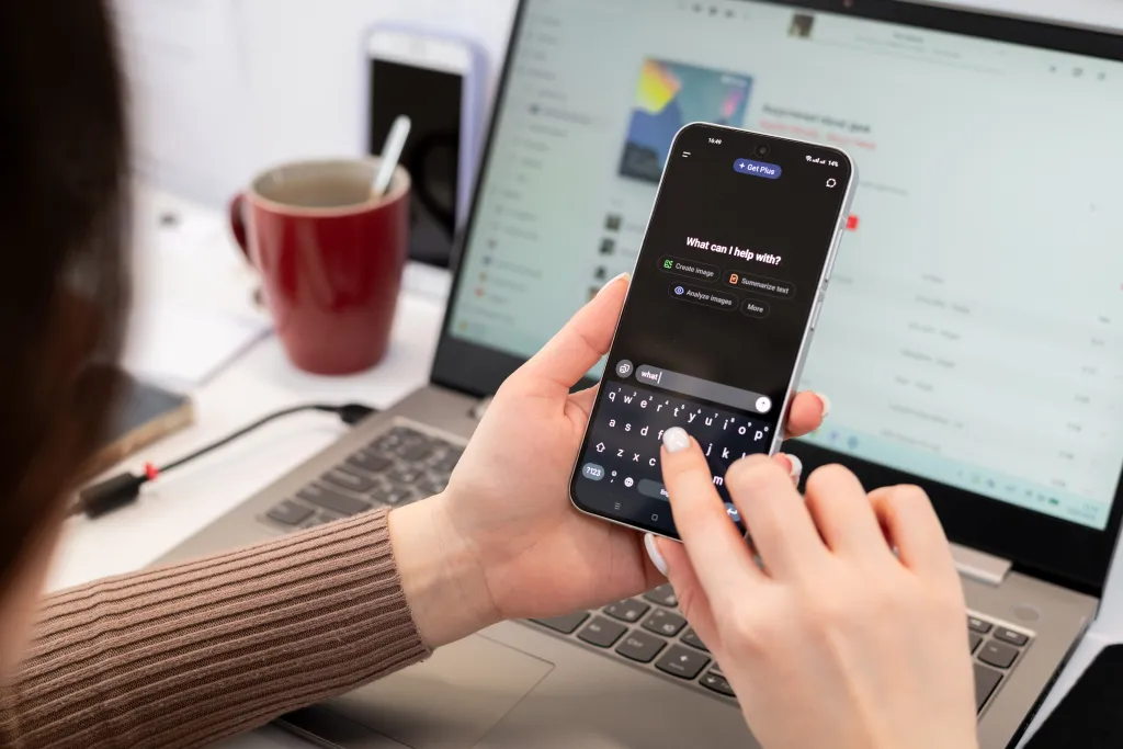 Woman using a smartphone with ChatGPT AI assistant app in a modern workspace with a laptop and coffee.