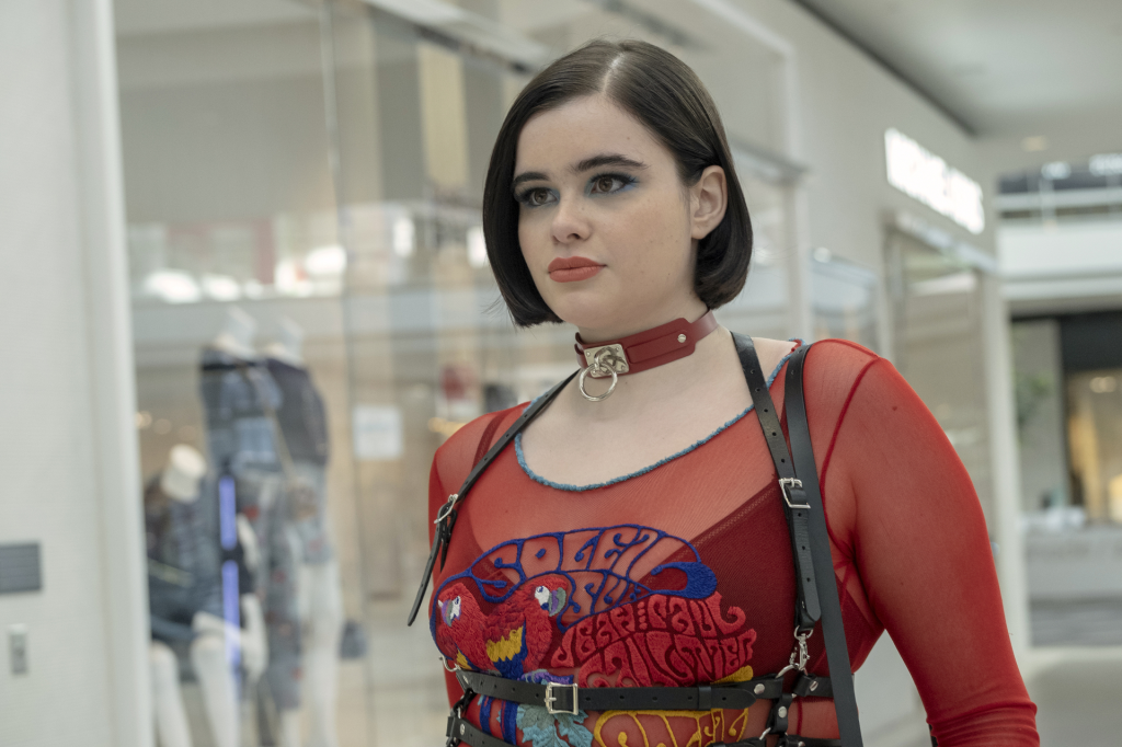 A young woman in a sheer red top with embroidered parrots and a leather harness.