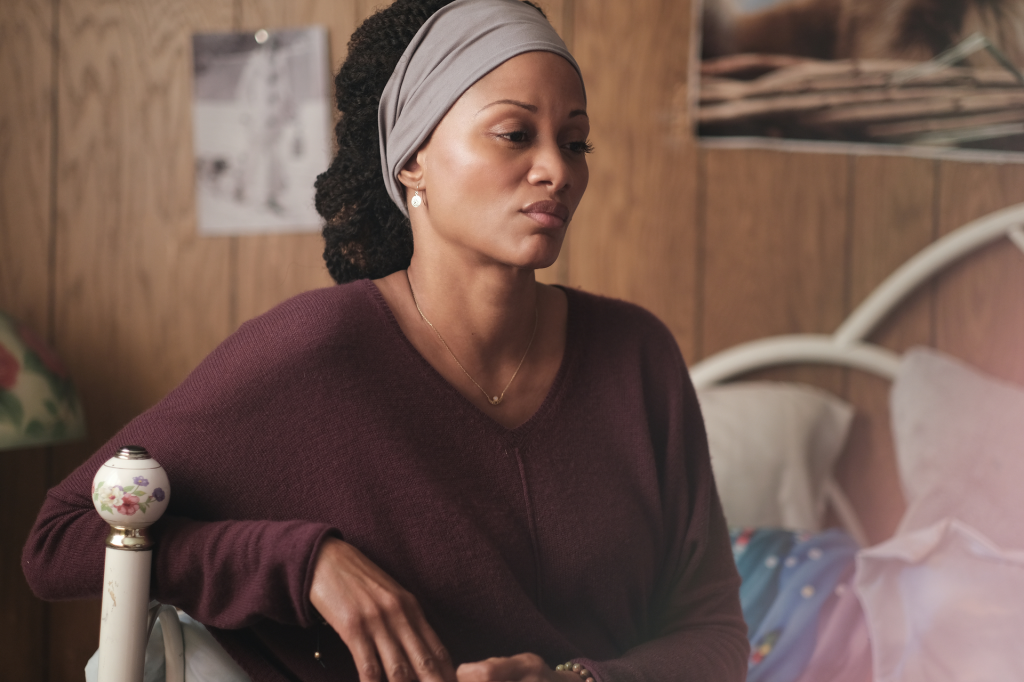 A woman with dark curly hair tied back with a grey headband wears a maroon V-neck sweater and looks thoughtfully into the distance.