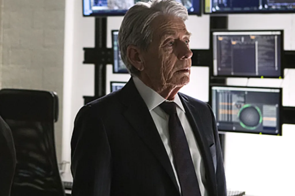 An older man in a suit and tie standing in front of multiple computer monitors.