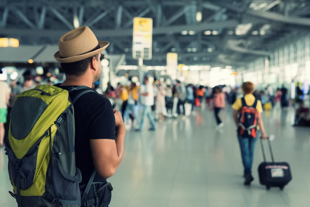 A man with a straw hat and green backpack looking at other travelers in an airport terminal.
