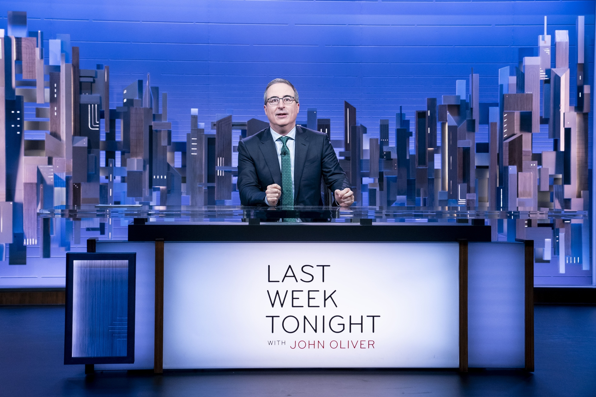 John Oliver standing at a desk with "Last Week Tonight with John Oliver" written on it, against a backdrop of a stylized cityscape.