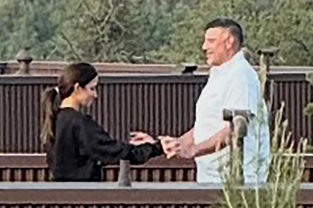 Two people, a woman and a man, standing and holding hands over a railing with greenery in the background.