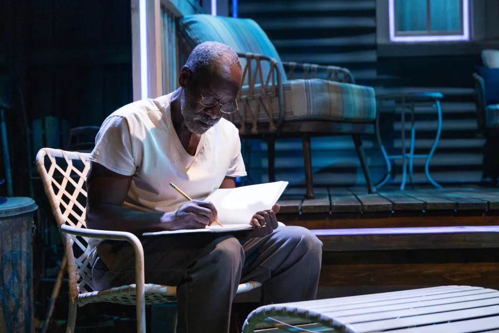 A man in glasses sketching in a notebook, seated on a white patio chair