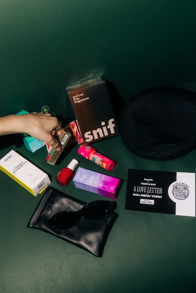 A hand reaching for a bottle on a table with a variety of beauty products, sunglasses, a hat, and an event invitation.