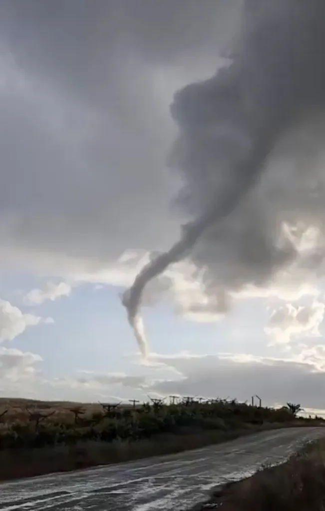 A funnel cloud extending from dark storm clouds over a road and dry land.