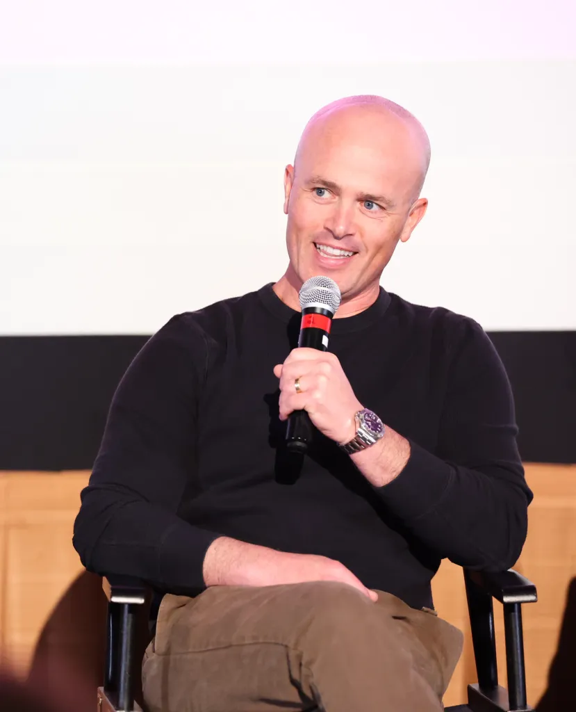 Chad Feehan wearing a black long-sleeve shirt and brown pants, smiles while holding a microphone.