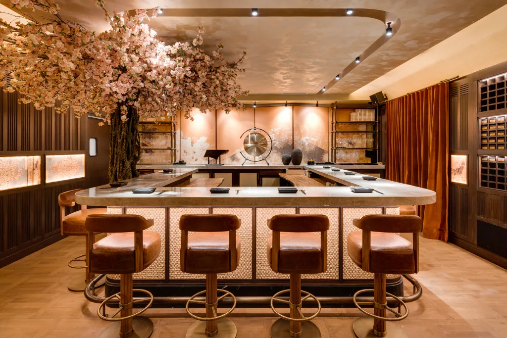 Interior of a restaurant with a central marble bar, brown leather stools, and a large artificial cherry blossom tree.