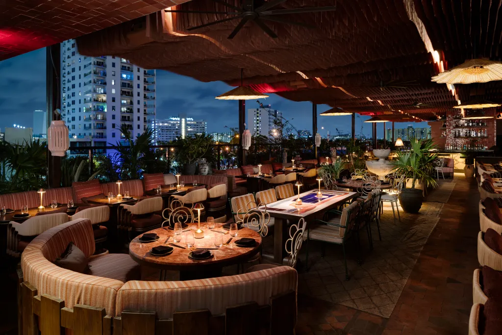 An outdoor rooftop restaurant with various seating arrangements, illuminated by warm lighting, against a backdrop of a city skyline at dusk.