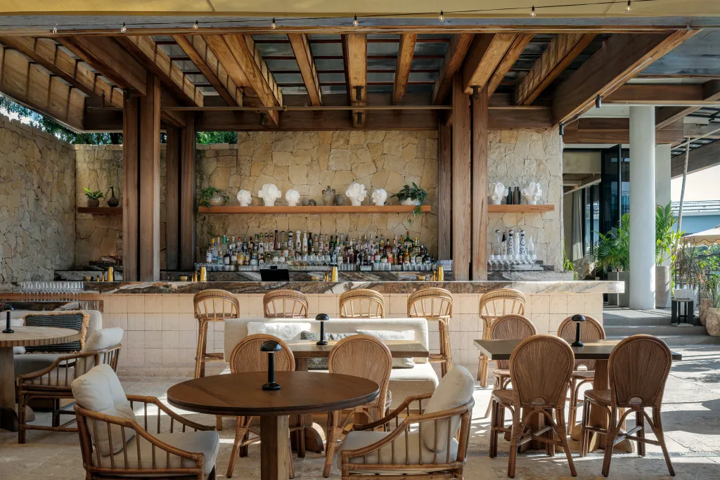 Architectural photography of a bar area with wooden beams, a stone wall, and rattan furniture in the Mila restaurant in Miami.