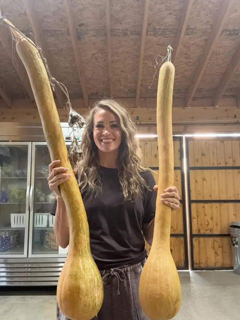 A woman smiling while holding two long, pale yellow squashes in front of her.