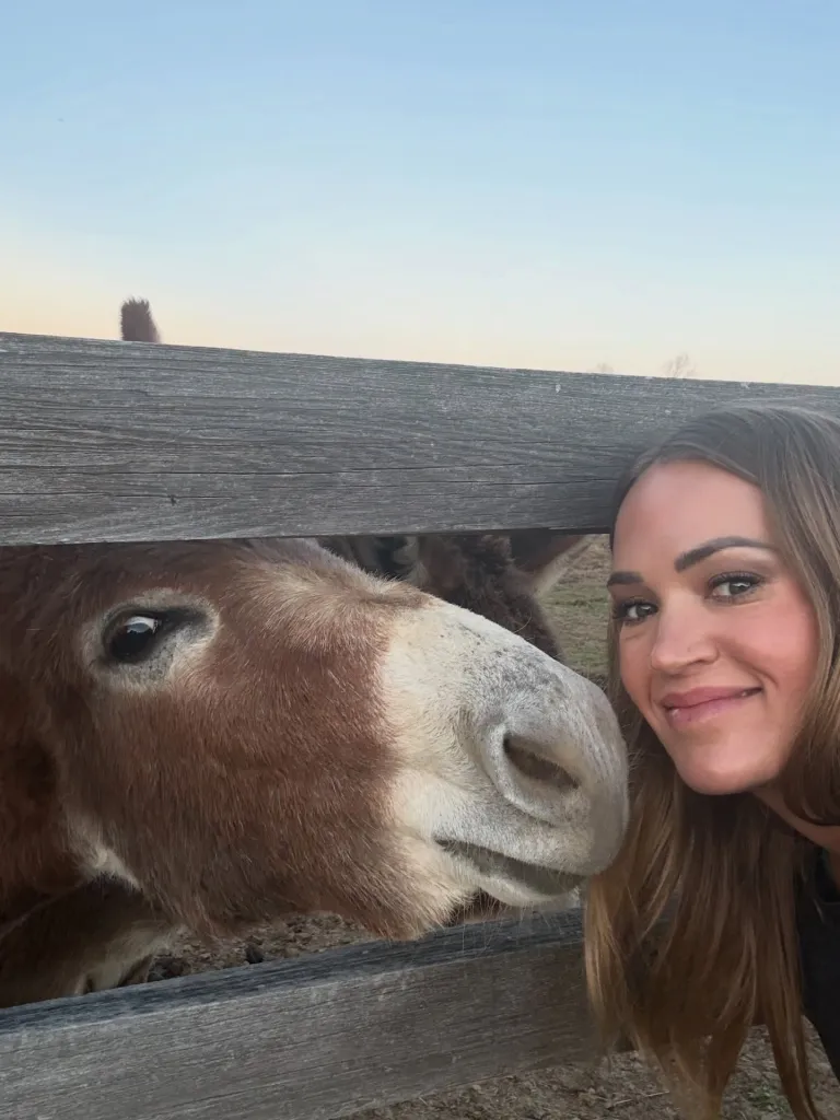 Carrie Underwood smiling next to a donkey at a wooden fence.