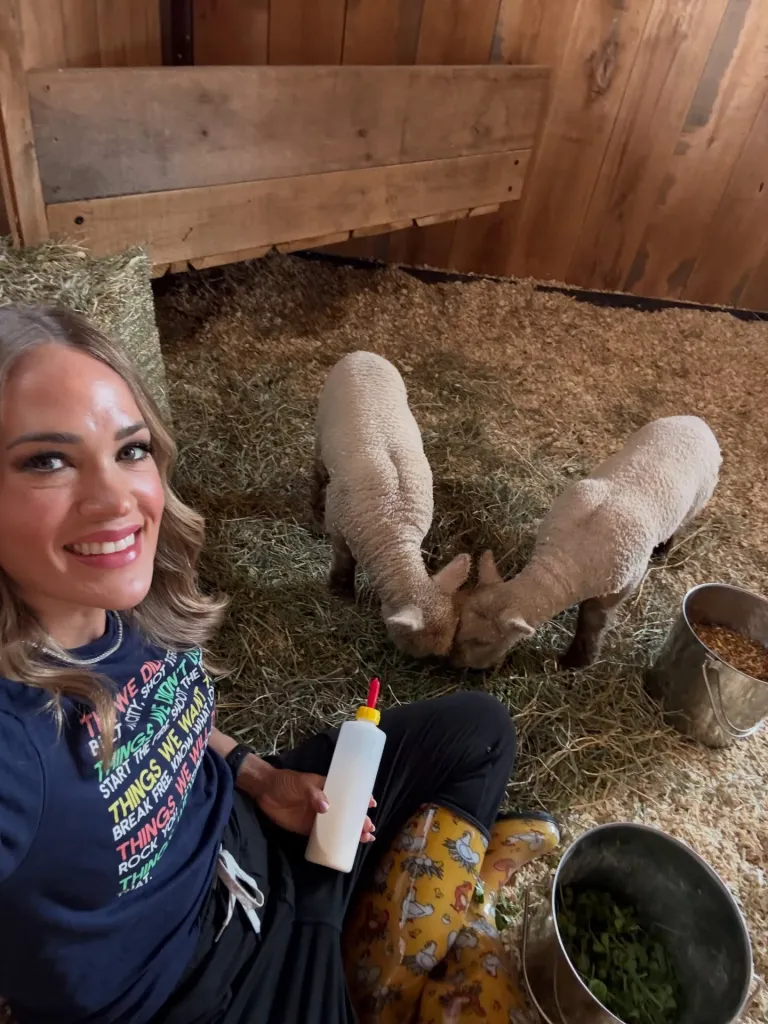 Woman sitting in a barn, holding a milk bottle and smiling, with two lambs and buckets of feed nearby.